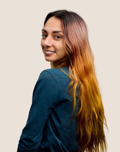Retrato de una persona del equipo de Apple Retail sonriendo con cabello largo café mirando por encima del hombro a la cámara.