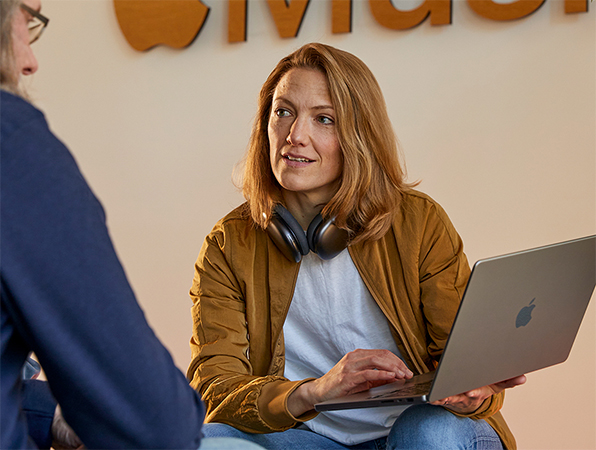 Dwie osoby z zespołu Sales and Business Development firmy Apple podczas rozmowy. Jedna osoba trzyma MacBooka i siedzi naprzeciwko ściany z logo Apple Music.