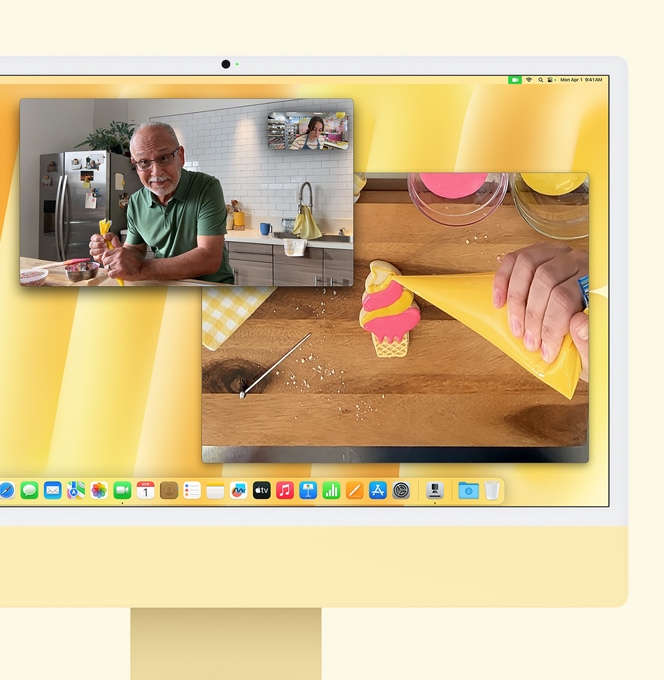 iMac Galben. Demonstrație a funcției Vizualizare birou. Pe ecran apare un brutar într-un apel video cu altă persoană. Pe un ecran apar brutarul și fața bărbatului. Pe celălalt ecran se văd mâinile brutarului care glazurează un fursec