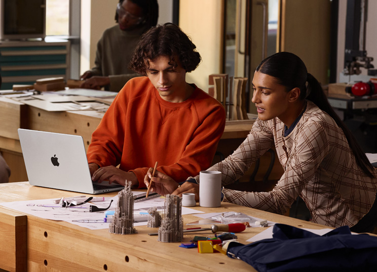 Zwei Personen arbeiten an einem Tisch gemeinsam an einem Projekt, eine Person tippt auf einem MacBook, die andere benutzt einen Stift und ein Lineal auf Papier, sie sind umgeben von Kunstbedarf.