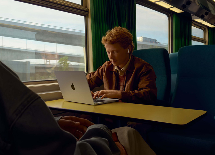 Eine Person, die AirPods trägt, sitzt an einem Fensterplatz im Zug und arbeitet an einem MacBook.