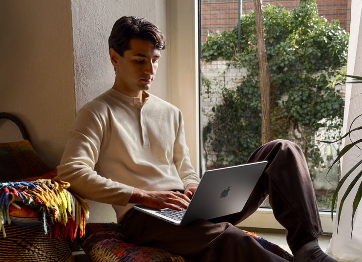 Eine Person, die ein MacBook benutzt, sitzt neben einem Fenster auf dem Boden.