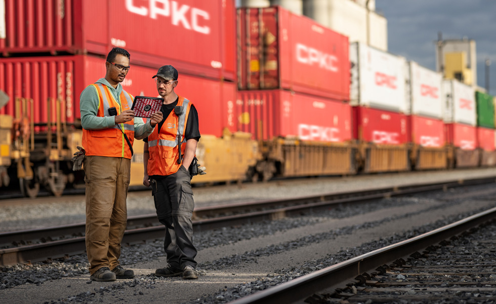 Duas pessoas da equipe da ferrovia consultam um iPad no pátio ferroviário, ao lado de um trem parado carregado de contêineres