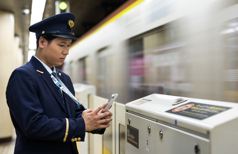 一名站務人員站在月台上使用 iPhone 掃描行動條碼，一班列車正行駛通過。
