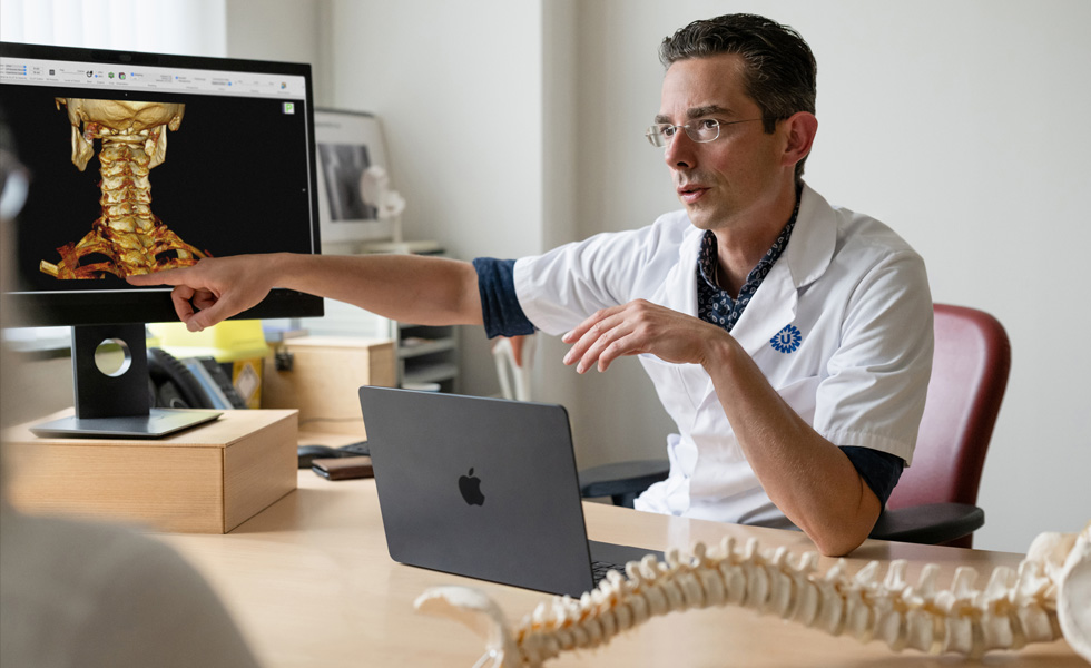 En medisinsk fagperson peker på en 3D-gjengivelse av skjelettet i en nakke, vist på Studio Display, mens en MacBook står på skrivebordet