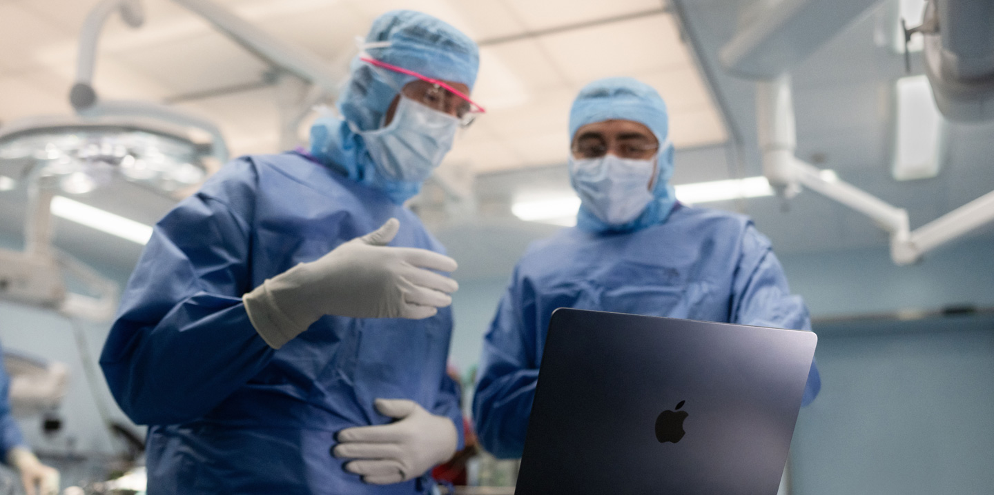 To kirurger i et operasjonsrom ser på en MacBook, og den ene gestikulerer mens den andre ser på