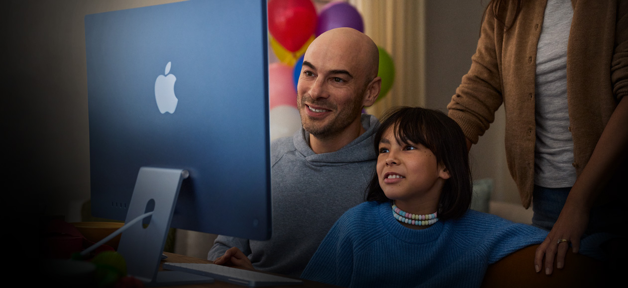 Dua orang duduk bersama menggunakan iMac, sementara orang ketiga berdiri di belakang mereka untuk mengamati.