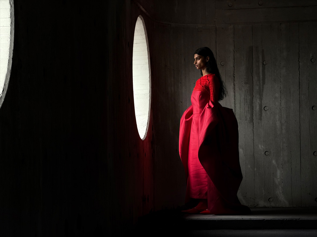 Una mujer con un largo vestido rojo de pie en medio de una habitación vacía mientras la fotografían. La distancia focal de la imagen se va alternando en la app Fotos para mostrar las capacidades del sistema de cámaras Pro: 100 mm